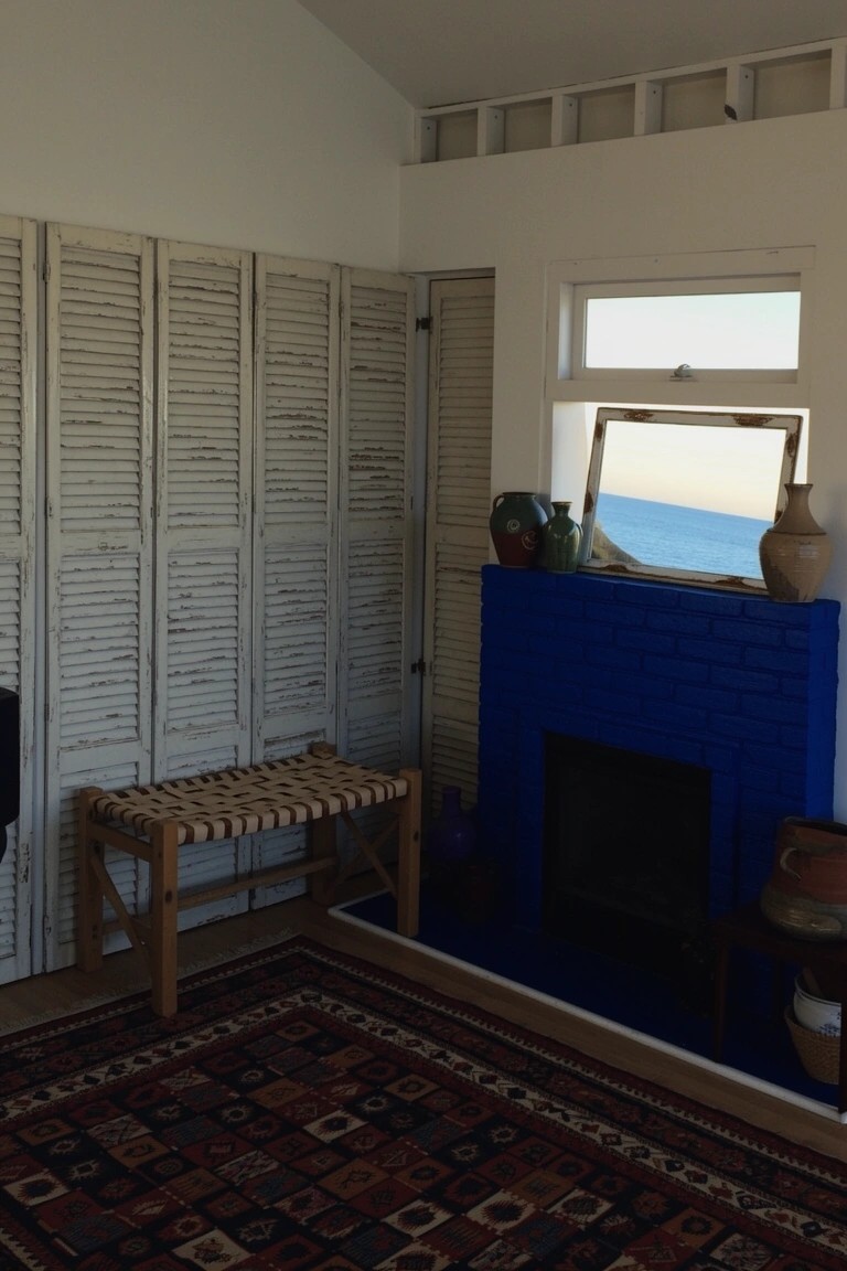 Surf house living room corner featuring a vibrant blue brick fireplace, white louvered screens, wooden bench, and pottery with ocean view through window