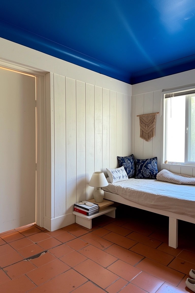 Surf house room featuring a vibrant blue ceiling, white paneled walls, low platform bed with pillows, macrame hanging, and red tile floor