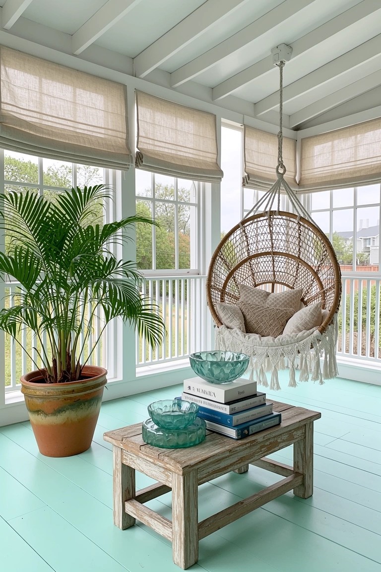Coastal sunroom featuring a hanging rattan swing chair with pillows, wooden table holding turquoise glass bowls and books, potted palm plant, mint green floor, and beige curtains on white-framed windows