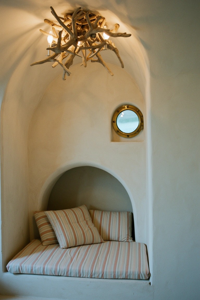 Arched wall alcove with cushioned bench seat, striped pillows, driftwood chandelier above, and brass porthole window in smooth white niche