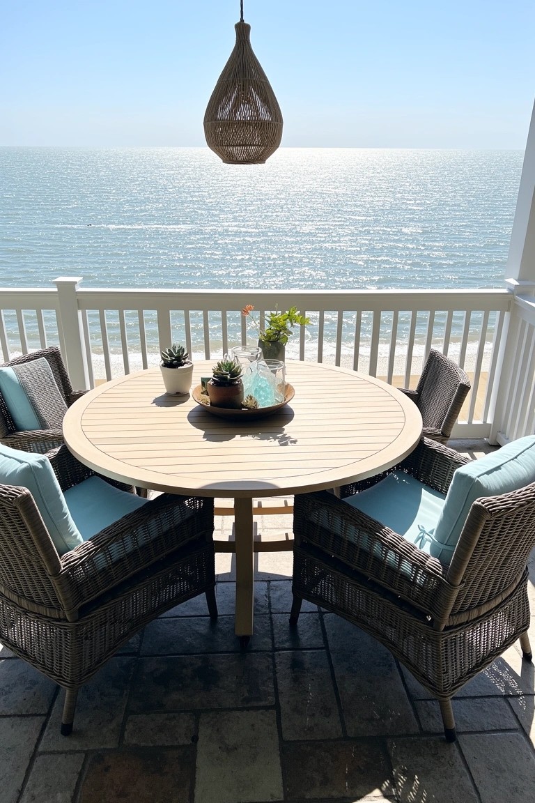 Round wood dining table with aqua wicker chairs and potted plants on balcony deck facing ocean