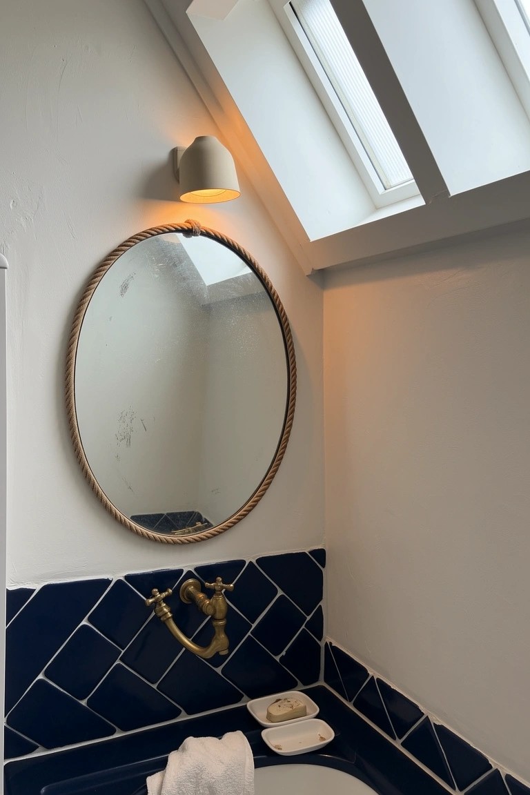 Round mirror edged with rope above blue tiled bathtub and brass faucet in beachy bathroom
