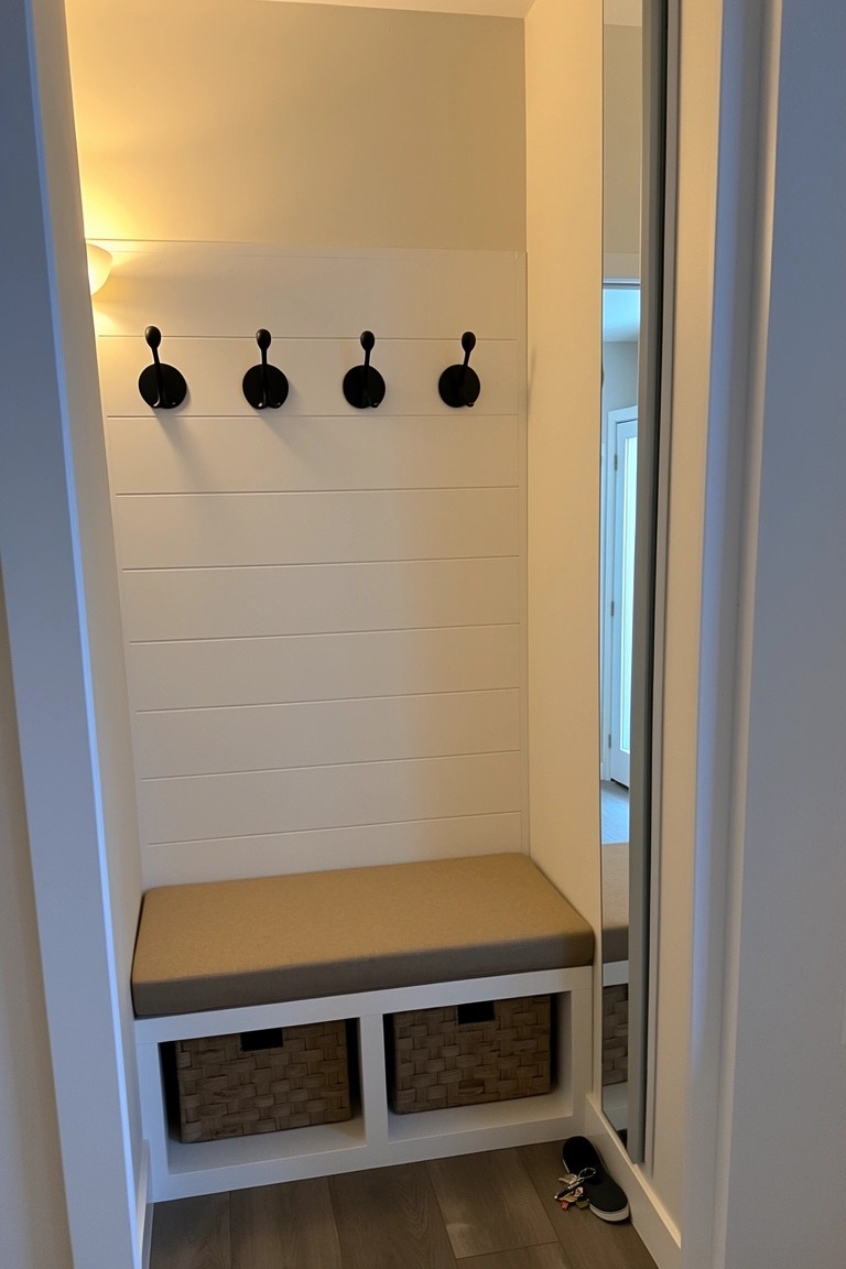 Narrow white shiplap entry nook with wooden wall hooks above a beige cushioned bench and wicker storage baskets