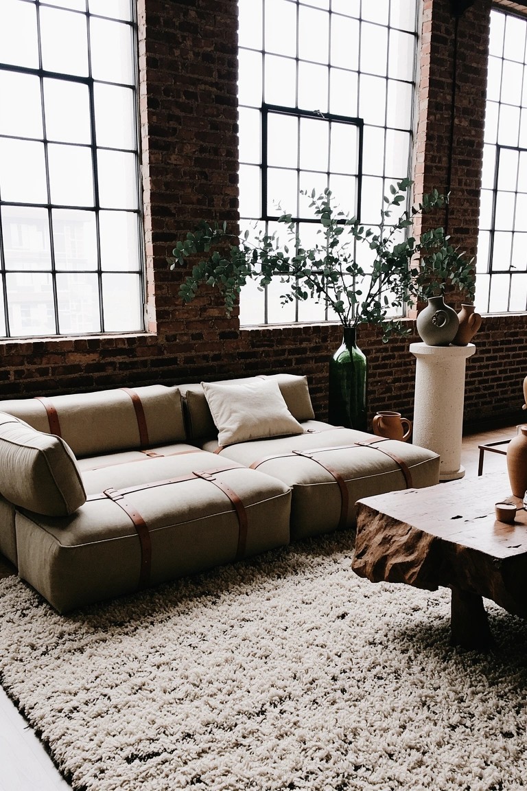 Beige modular sofa with tan leather straps on a white shaggy rug in a brick-walled loft living room with plants and pottery