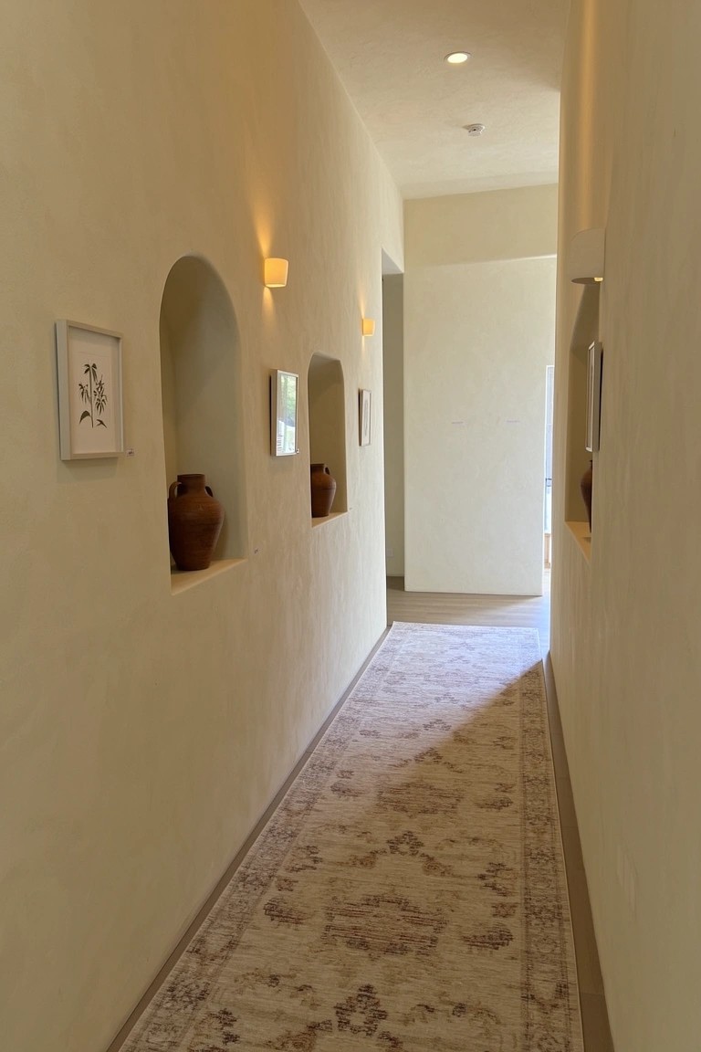 Beige stucco hallway with recessed wall niches holding terracotta pots and small frames, wall sconces, and a neutral runner rug leading to a door