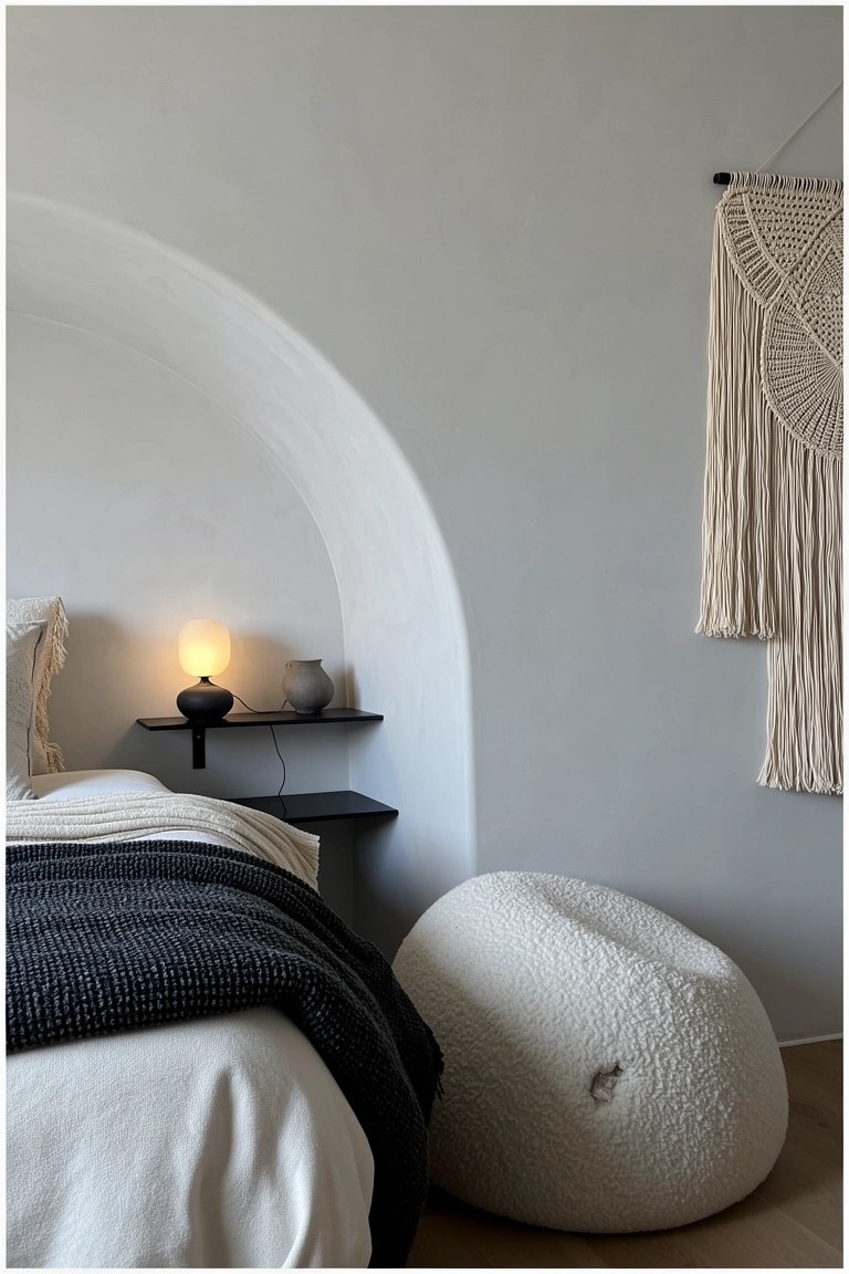 Cozy modern boho bedroom with curved white wall niche framing the bed, black shelf lamp, macrame hanging, and white pouf stool