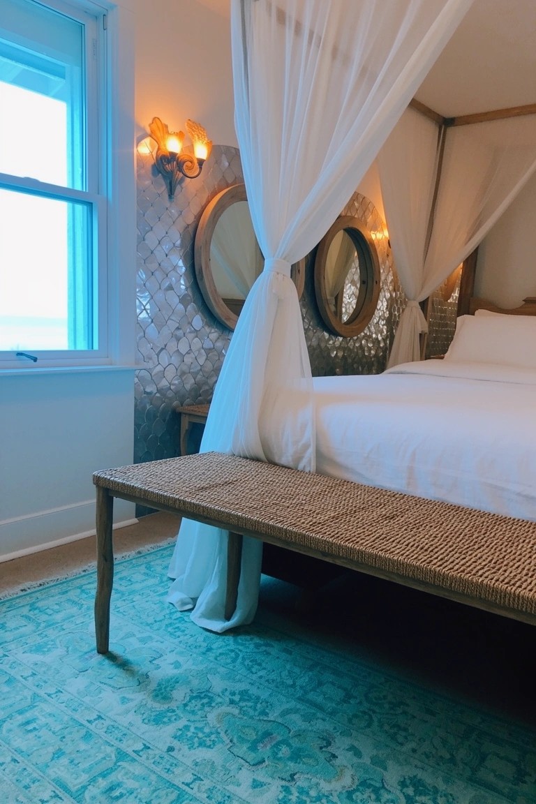 Bedroom with canopy bed draped in white sheers against iridescent scalloped tile wall with round mirrors, turquoise area rug, and rattan bench at bed's foot