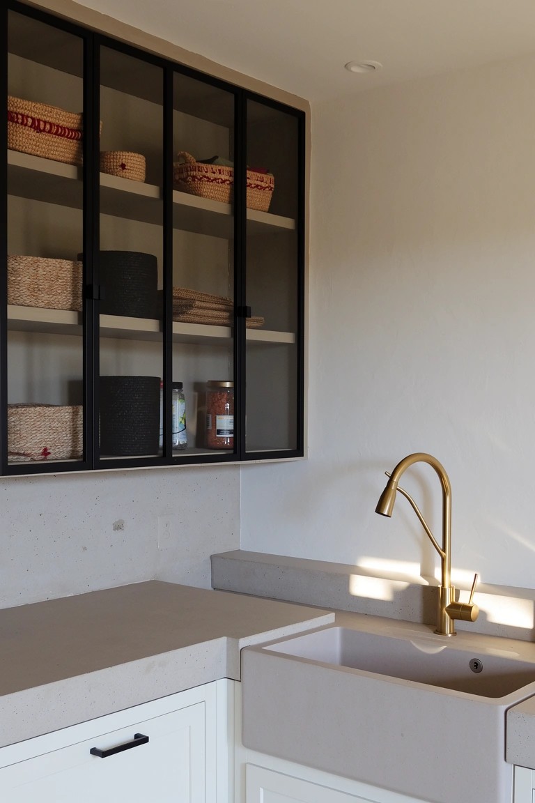 Bright kitchen corner with black metal-framed glass cabinets displaying woven baskets, jars, and containers on open shelves, white sink with gold faucet, and light gray counters