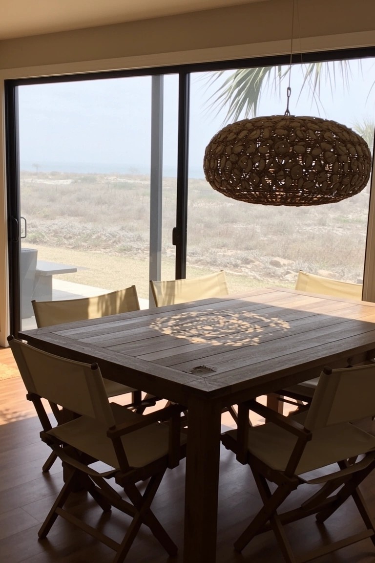 Casual beach dining room with open sliding doors to ocean view, wooden table, beige folding chairs, and woven pendant light