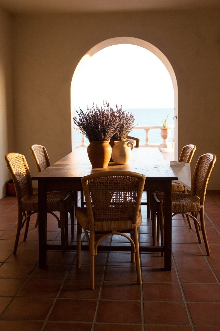 Casual beach dining room with wicker chairs around a wooden table on terracotta floors, pottery vases of lavender, and arched window to the sea