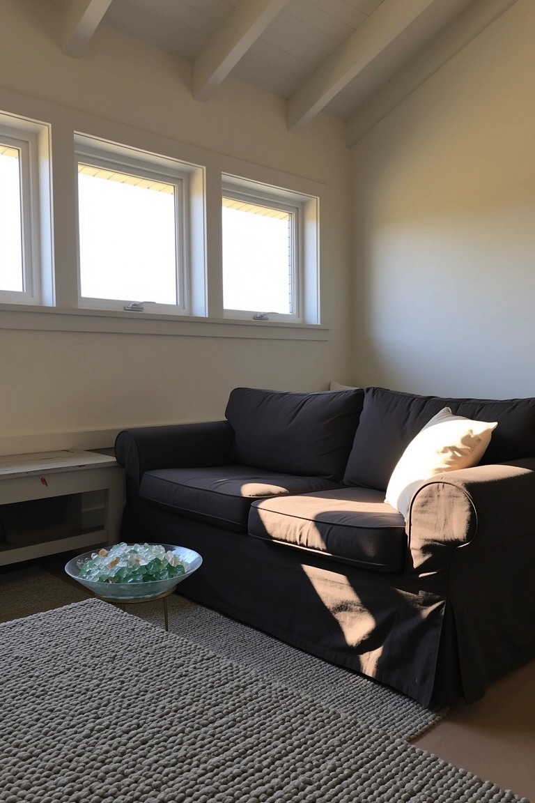 Light-filled attic nook with exposed wooden beams, dark gray sofa facing triple windows, side table holding sea glass bowl, neutral rug on wood floor