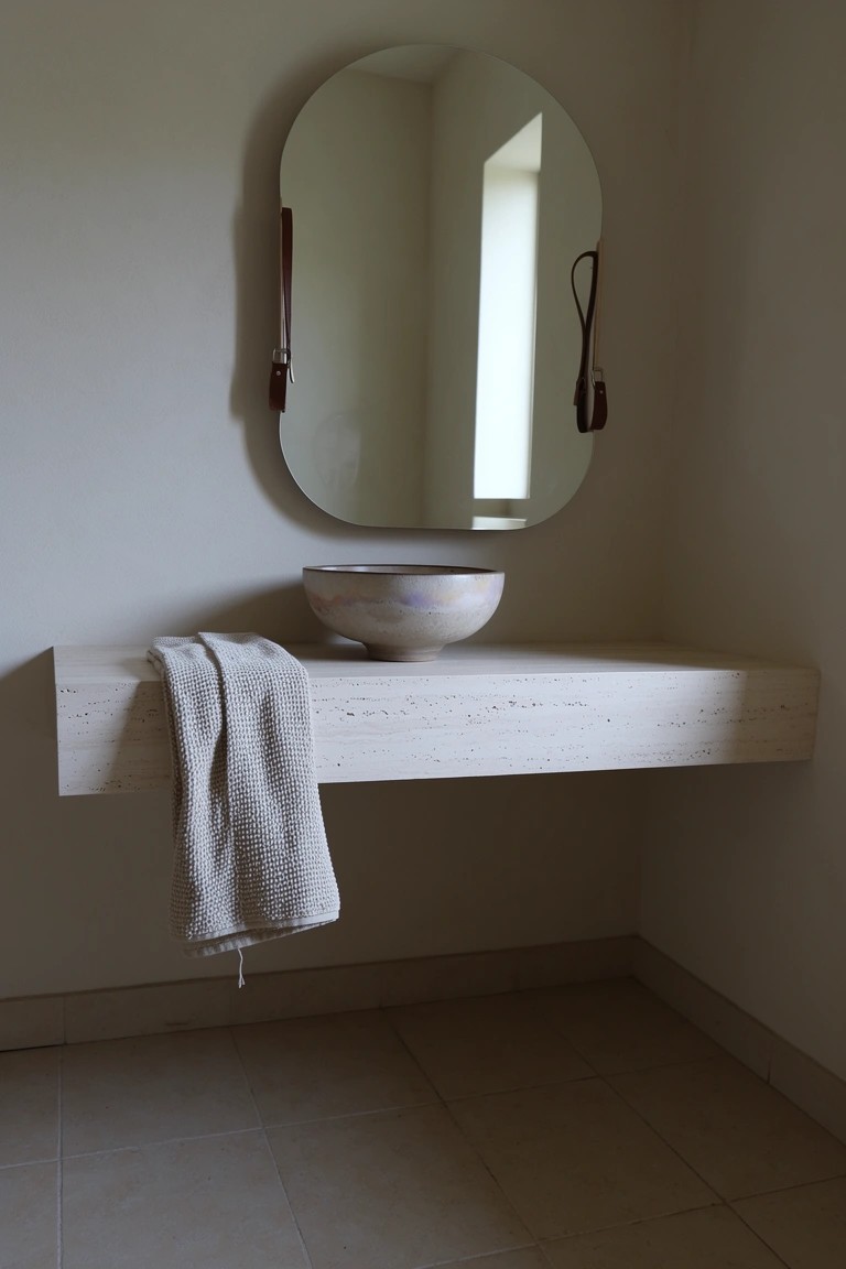 Floating white shelf holding a handmade ceramic basin sink, beige towel, and oval mirror with leather strap in a neutral bathroom
