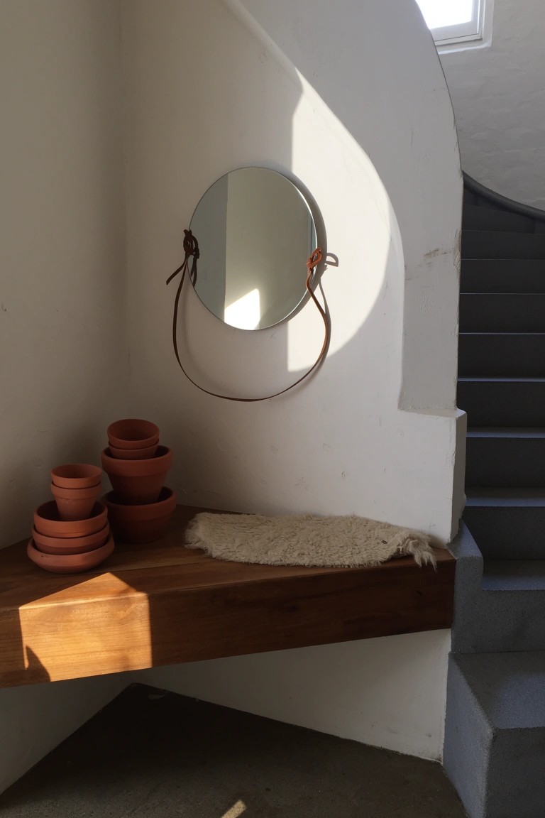Wood shelf in white curved corner holding stacked terracotta pots and draped sheepskin, with round leather-strapped mirror above near stairs