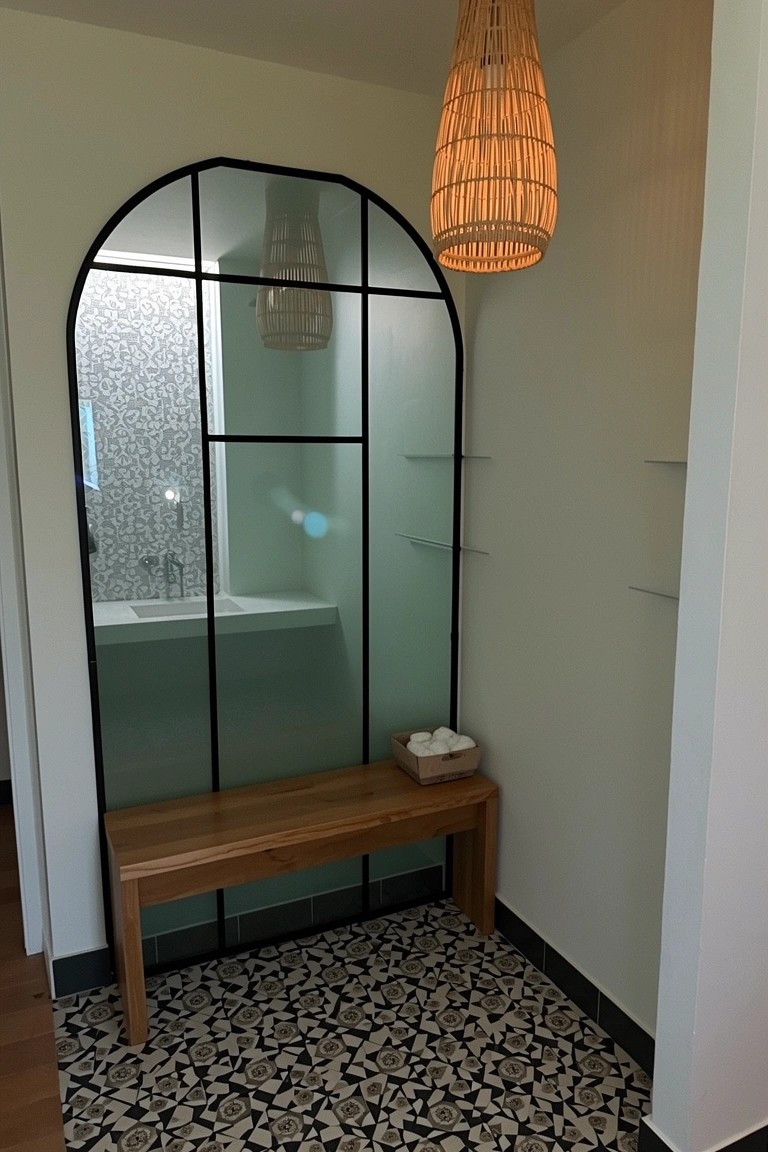 Arched black-framed mirror reflecting a bathroom sink with wooden bench, shelves, rattan pendant light, and patterned tile floor below