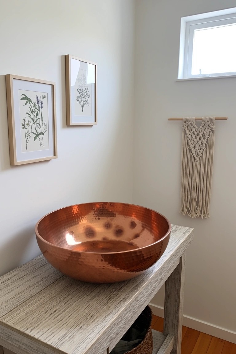 Bathroom corner featuring a large hammered copper bowl sink on a light wood vanity table, with botanical prints on the wall and a macrame hanger nearby