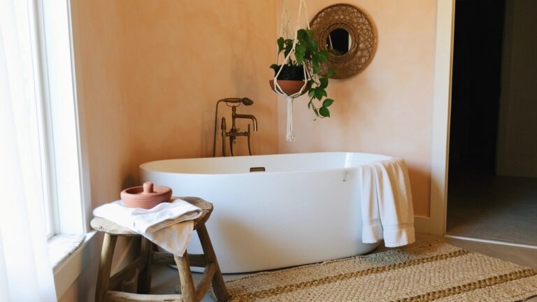 Boho bathroom with soft terracotta walls, white freestanding tub, macrame hanging plant, and wooden stool with towels