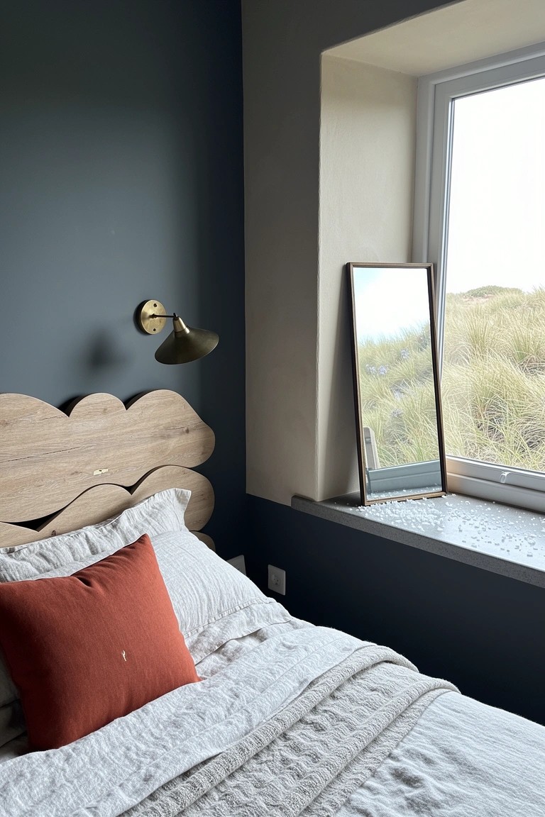 Beachy bedroom with tall leaning mirror by window reflecting dunes, dark teal wall, wavy wooden headboard, brass sconce, and light linens with orange pillow