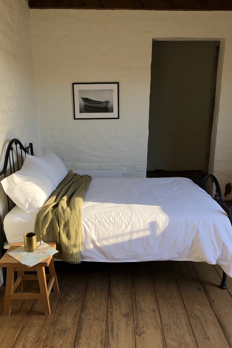 Simple bedroom featuring black iron bed frame with white bedding and green throw, wooden stool side table holding mug and booklet, black-and-white boat photo on white plaster wall, green door, wooden floors