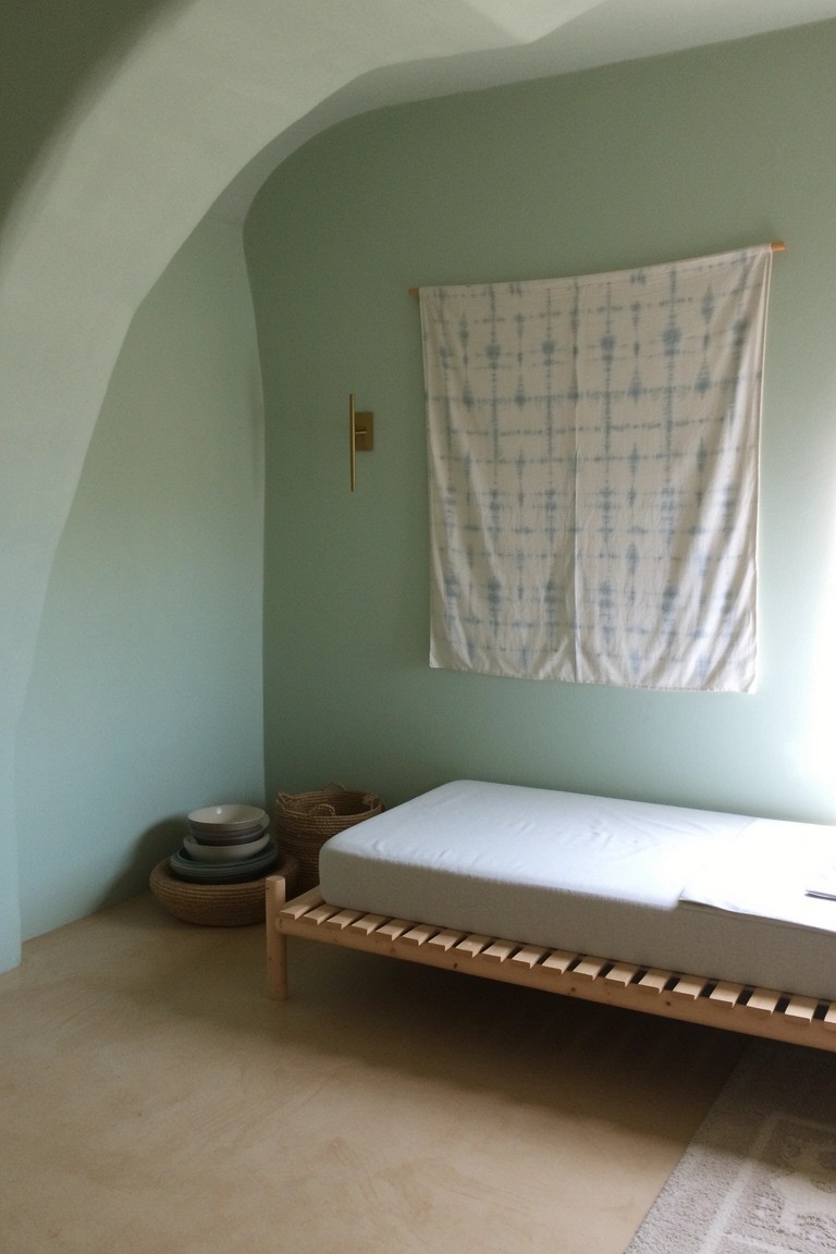 Sage green curved bedroom wall with low wooden platform bed, hanging indigo textile, and woven basket