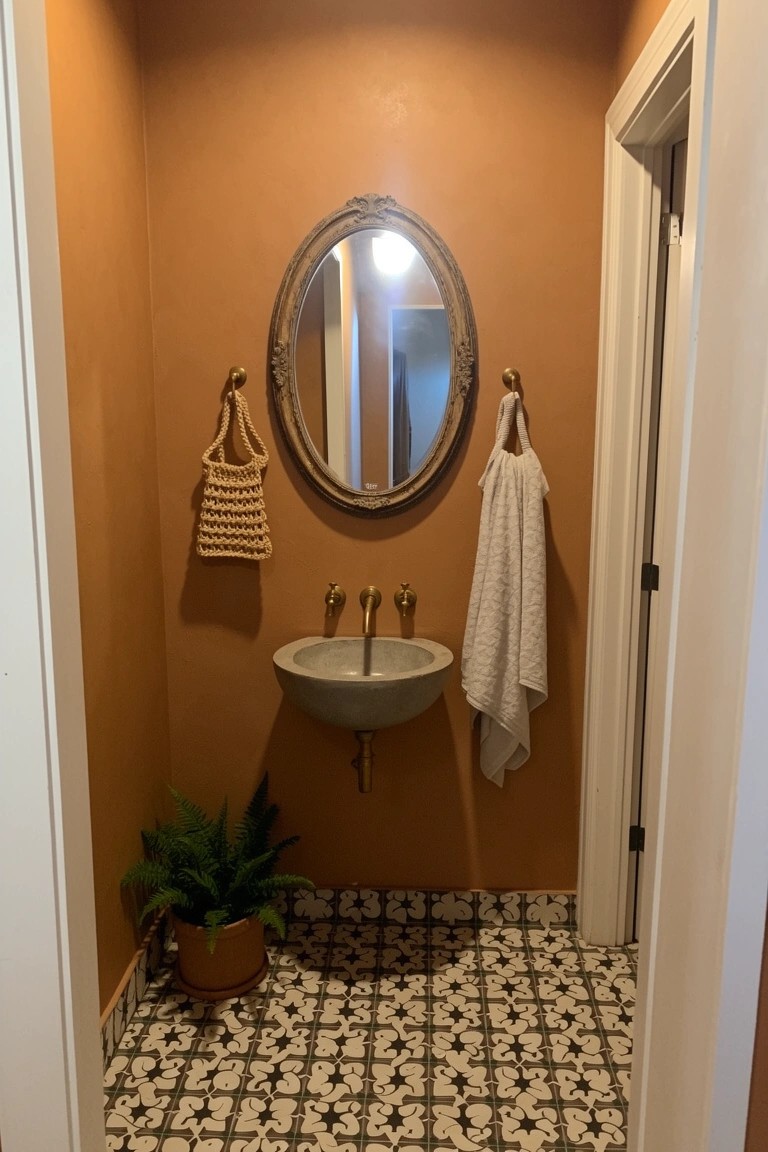 Small powder room with warm terracotta walls, oval vintage mirror holding a woven basket and towel, potted fern, white wall-mounted sink, and black-and-white patterned tile floor
