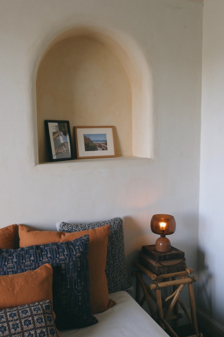 Bedroom corner showing arched wall niche with two framed photos, terracotta lamp on rattan side table next to bed layered with orange and blue pillows