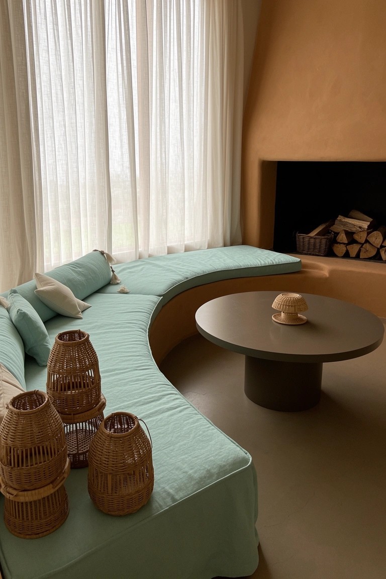 Curved teal banquette sofa in terracotta-walled living room with sheer curtains, round table, woven baskets, and fireplace nook