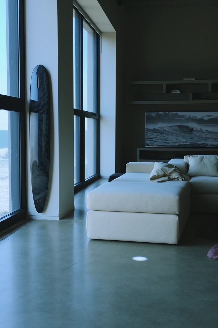 Minimalist living room with black surfboard leaning against floor-to-ceiling windows next to white sofa and ottoman