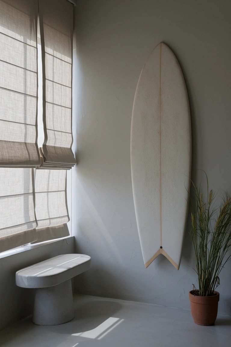 White surfboard hanging vertically on light gray wall next to window with beige blinds and simple stone bench in minimalist room corner