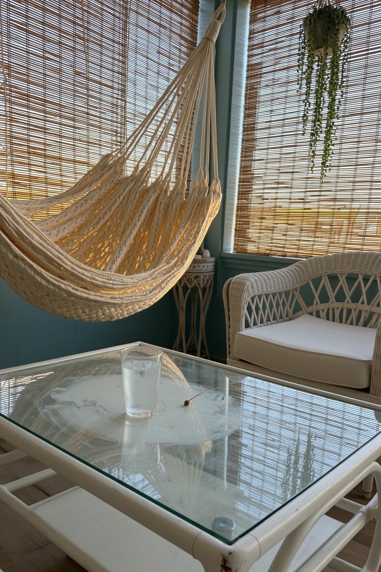 Beige woven hammock chair hanging in a coastal boho porch with bamboo blinds, wicker furniture, glass table and potted plants