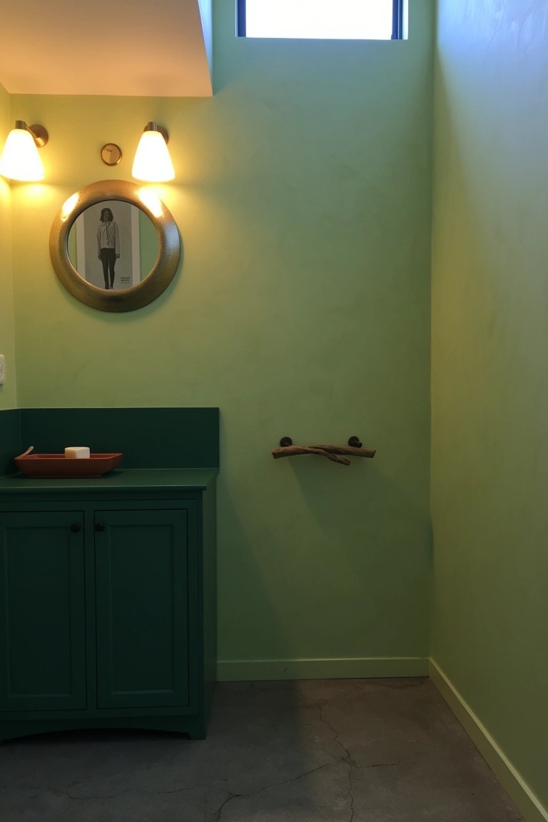 Compact powder room with pale green walls, dark green cabinet, round mirror, and natural branch towel bar