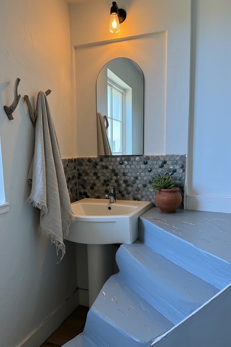 Cozy powder room under stairs with white corner sink, iridescent mosaic tile backsplash, arched mirror, potted succulent, and blue painted steps