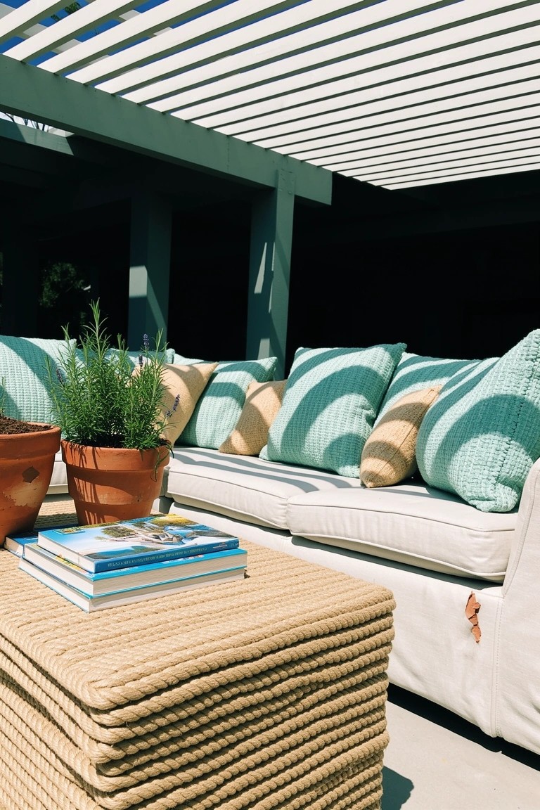 Outdoor patio lounge with white sofa, teal cushions, rope-wrapped wood coffee table holding books and plants, potted rosemary nearby