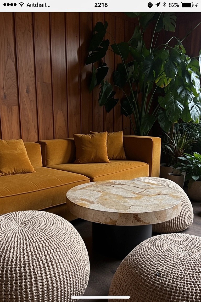 Warm wood paneled living room wall with mustard velvet sofa, round beige stone coffee table, cream woven poufs, and tall green plants creating an earthy boho nook