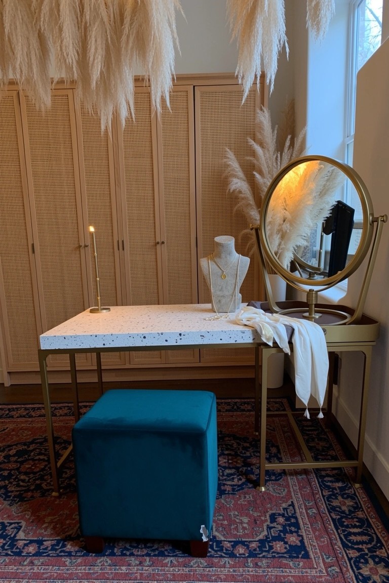 Boho chic vanity nook with hanging pampas grass, speckled white table on gold legs, turquoise velvet stool, cane-front cabinets, and gold mirror stand