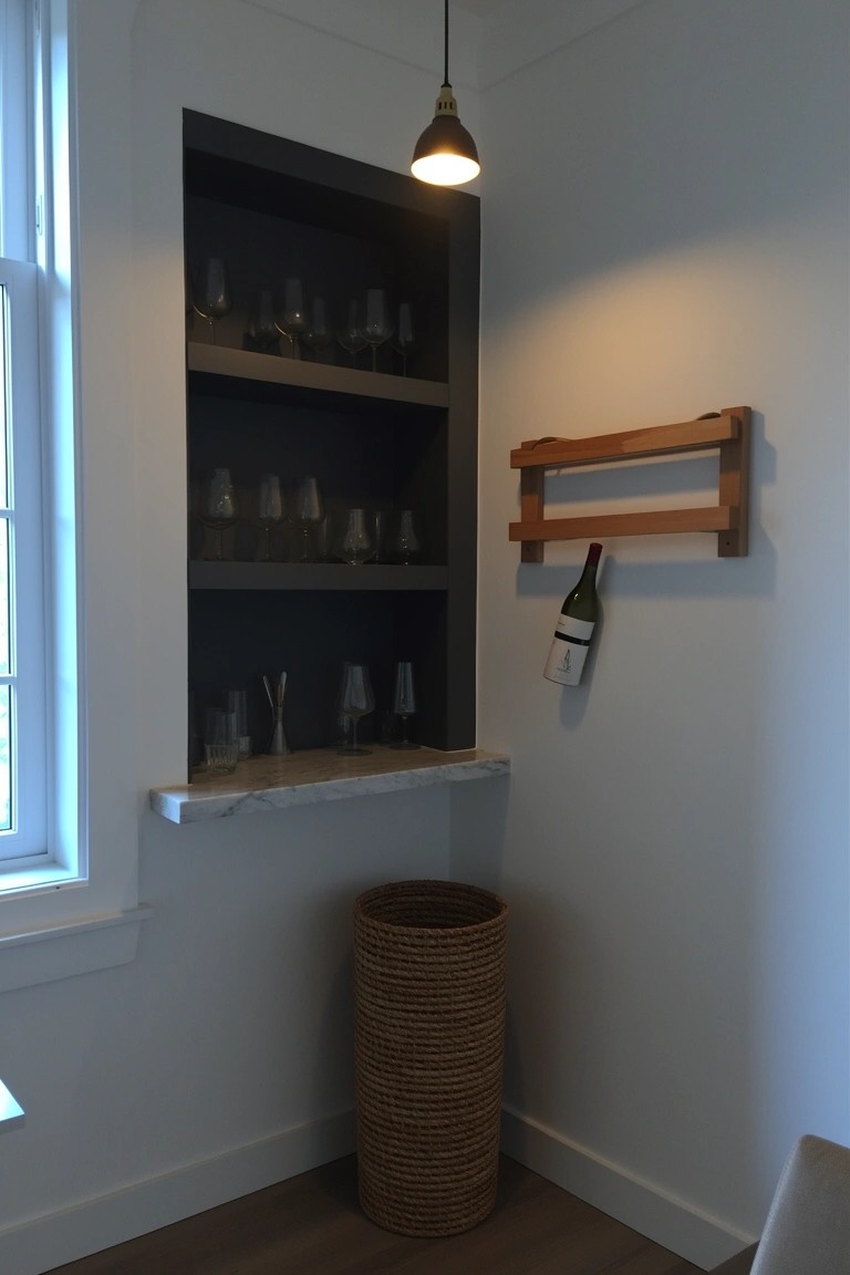 Recessed corner wall niche with dark shelves holding glassware, wooden rack with wine bottle and towel, and woven basket bin on wood floor