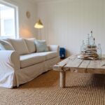 Cozy beach-style living corner featuring white shiplap walls, slipcovered white sofa, rustic wooden coffee table with clustered blue glass bottles, seagrass rug, brass floor lamp, and coral wall art