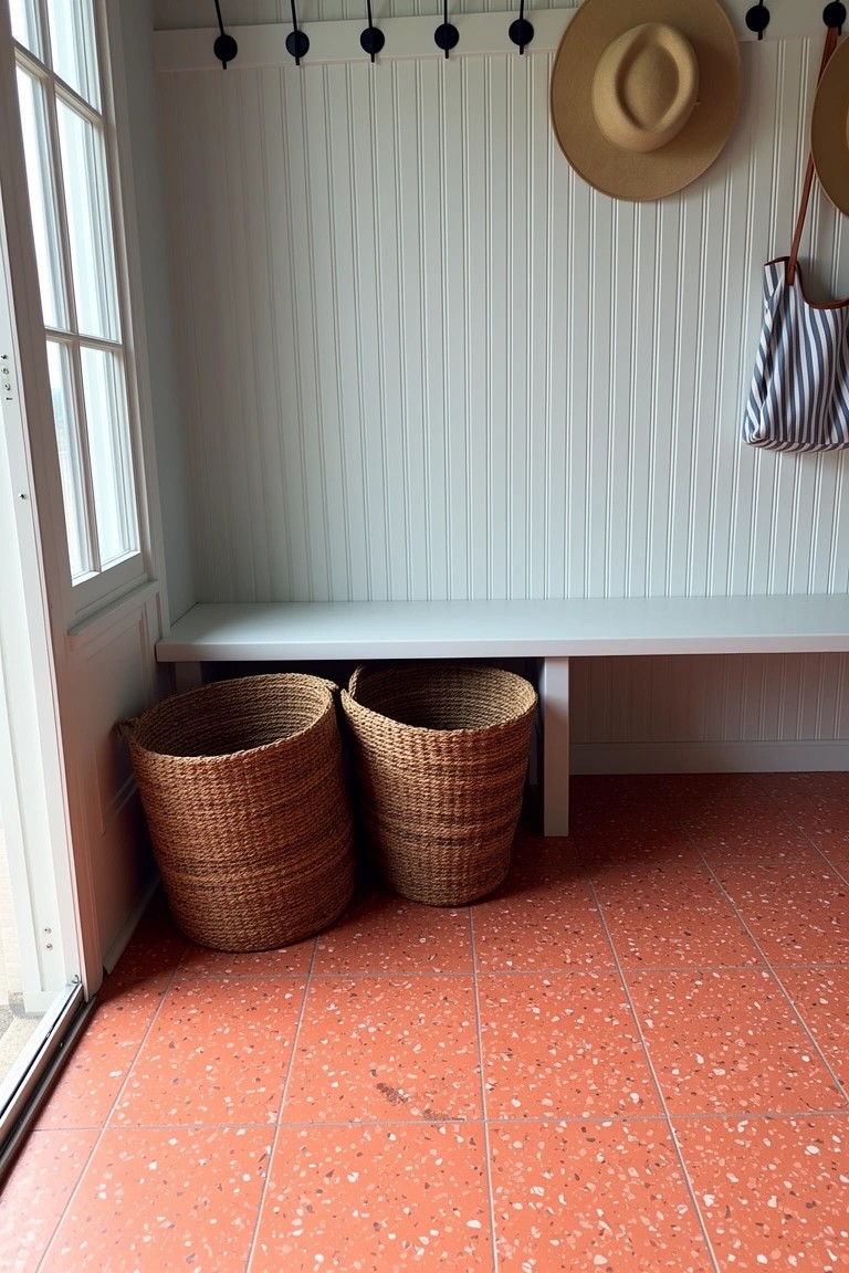 White wainscoted mudroom with wooden bench, two large woven storage baskets underneath, hats and bag on wall hooks, red terrazzo floor