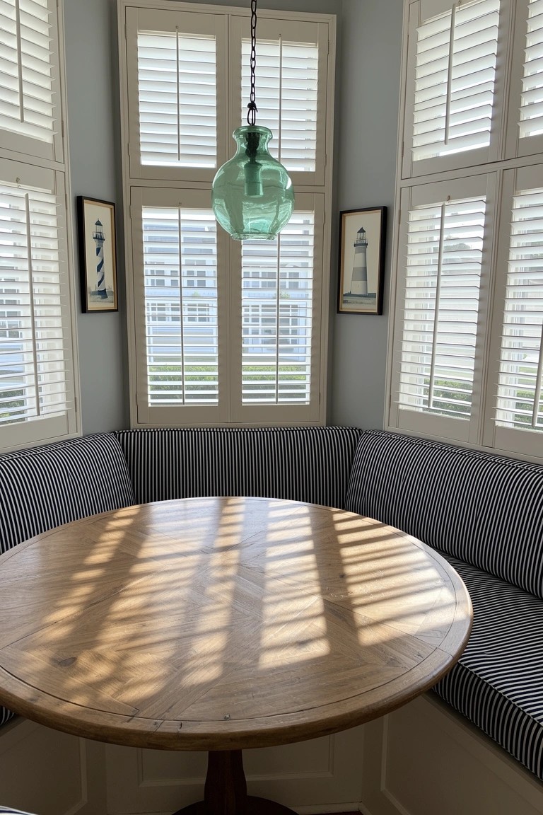 Bay window nook with round wooden table, striped banquette seating, green glass pendant light, and white plantation shutters