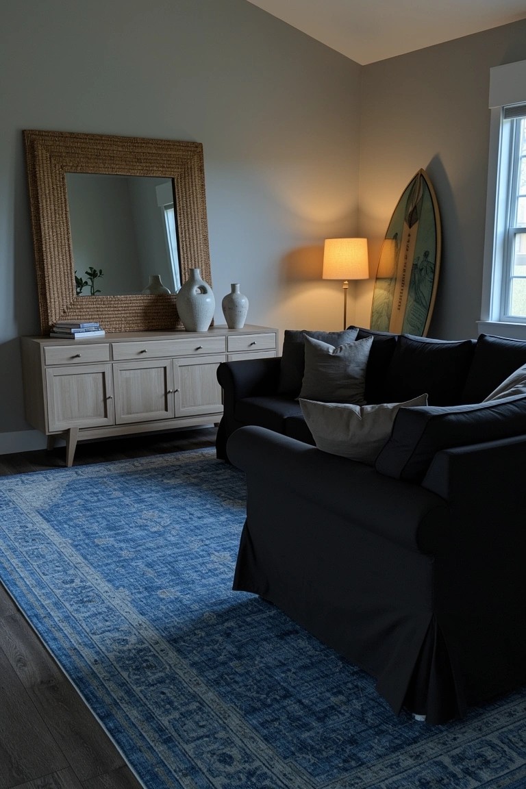Beach house living room corner featuring a black sofa on a blue rug, wooden credenza with vases, rattan mirror, lamp, and surfboard leaning by the window
