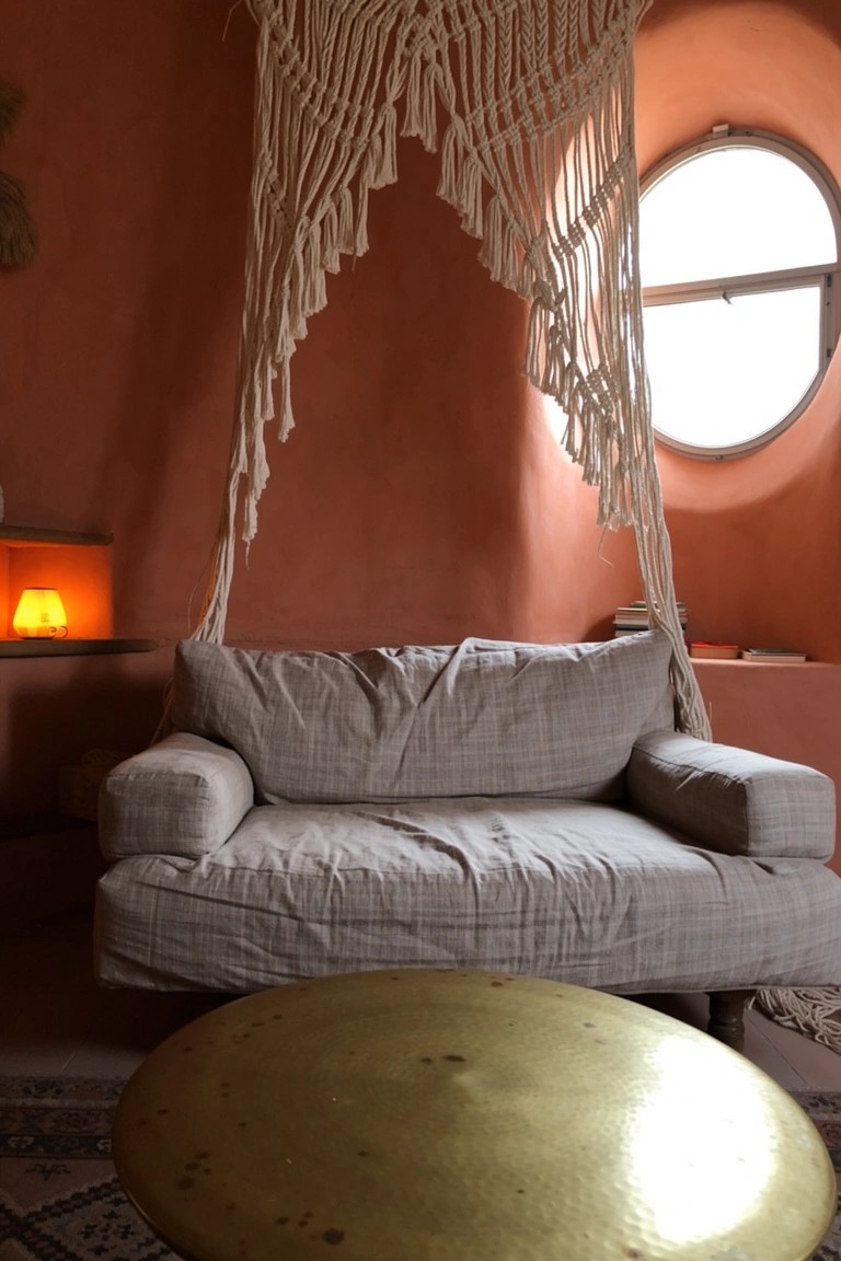 Boho seating nook with white macrame canopy draped over low gray sofa against terracotta wall, brass drum table, and small fire lamp