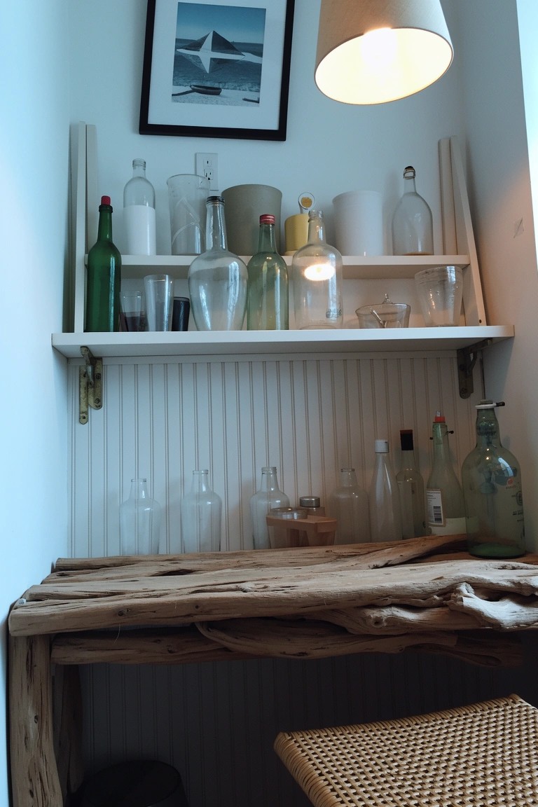 Open wooden shelves filled with colorful glass bottles, jars and white ceramics above a driftwood counter in a white beadboard nook