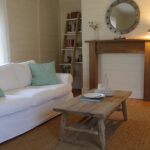 Cozy coastal living room with white slipcovered sofa, mint green pillows, seagrass rug, driftwood coffee table, shiplap walls, and round mirror over fireplace