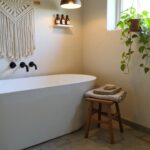 Bathroom corner with white freestanding tub, black faucet, hanging potted plant in macrame, wall shelf with bottles, and wooden stool holding towel