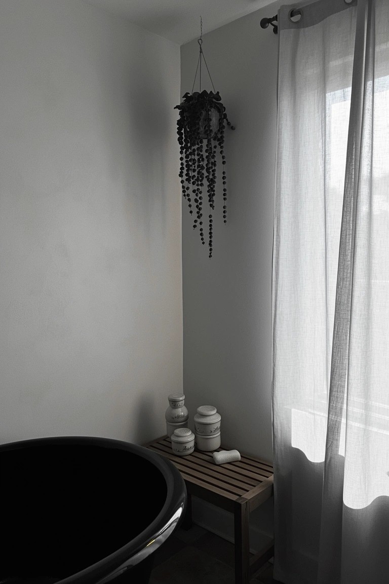Bathroom corner with black freestanding tub, trailing hanging plant, white jars on wood stool, and sheer curtains on window
