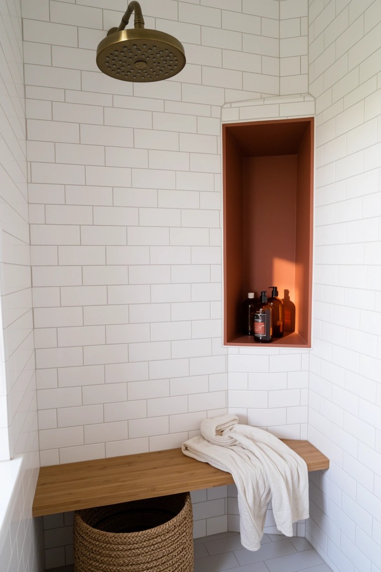 White subway tile shower corner with built-in wooden bench, draped white towel, terracotta wall niche holding amber bottles, rainfall showerhead above, and woven basket on floor