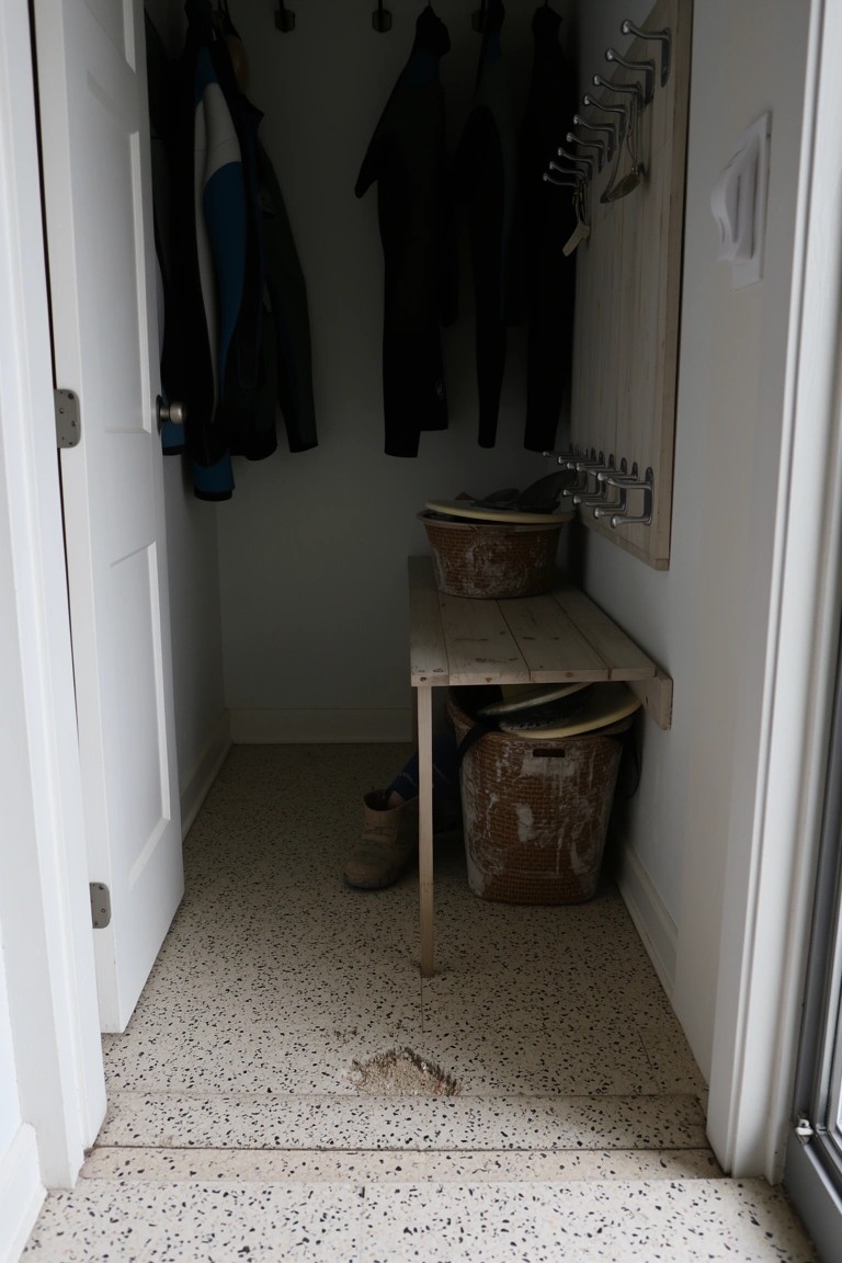 Narrow white entry closet with coats hanging on wall hooks and rail, wooden bench with woven baskets for shoe storage, tan shoes visible on speckled floor