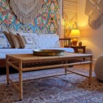Bohemian living space with vibrant floral wallpaper on one wall, low wooden daybed, potted plants in gold pot, woven pouf, and patterned rug on hardwood floors