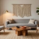 A light gray sofa sits in a white-walled living room with sheer curtains on a black-framed window overlooking trees and plants, a wooden coffee table with terracotta pots, large fiddle leaf fig in a basket, rattan armchair, macrame wall hanging, and gold arched floor lamp.