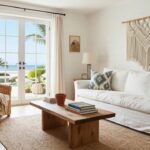 White slipcovered sofa with blue ikat pillow and throw on a low wooden coffee table with books and potted succulent, two rattan armchairs nearby, macrame wall hanging on shiplap wall, and French doors to ocean view in a light coastal living room.