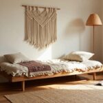 Light beige living room corner with wooden futon sofa covered in white linen cushions and a multicolored patterned throw, wooden shelves with baskets and books, potted plants, rattan floor lamp, macrame wall hanging, and wicker baskets on hardwood floor.